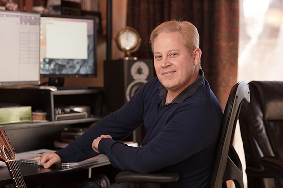 A smiling man with blonde hair sits in a black office chair in a home studio with computer monitors.