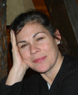 A woman with dark hair pulled back, resting her chin on her hand and smiling slightly.