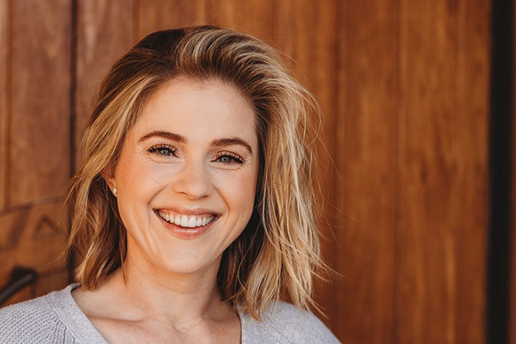 A smiling woman with blonde hair looking towards the camera against a wooden background.