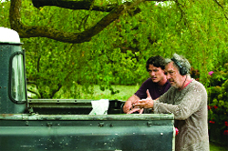 Two men stand by the bed of a dark pickup truck under the branches of a large green tree.