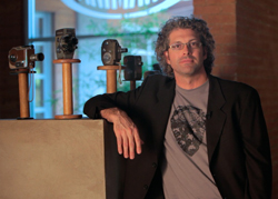A man with curly hair and glasses wearing a blazer leans on a pedestal next to several vintage film cameras.