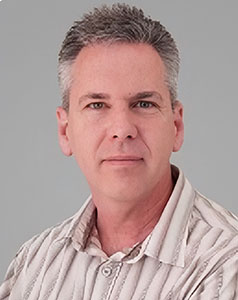 A man with short graying hair wearing a tan and white striped collared shirt against a gray background.