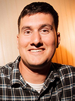 A smiling man with short dark hair wearing a black and white plaid shirt.