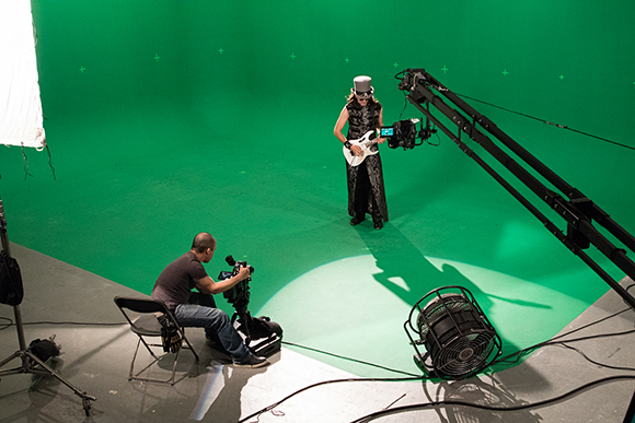 A woman in a black outfit plays a white guitar on a green screen while a camera operator films her.