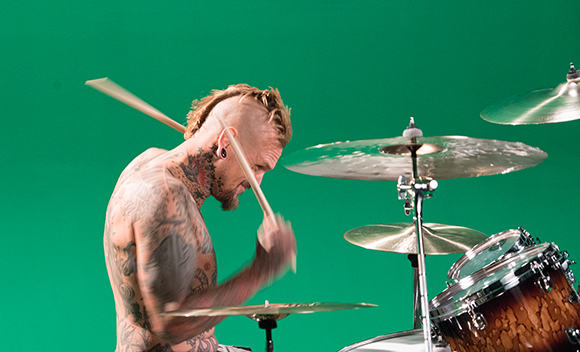 A shirtless, tattooed man with a mohawk plays a drum kit against a solid green backdrop.