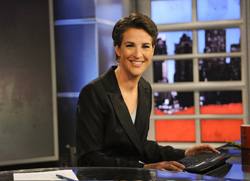 A smiling woman in a black blazer sits at a desk in a news studio.