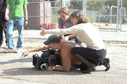 A camera operator lies on the ground filming while two crew members assist him outdoors.