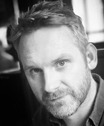 Black and white close-up portrait of a man with short hair and a beard looking toward the camera.