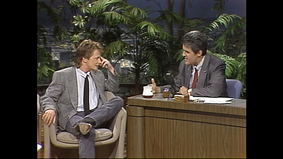 A man in a grey suit sits in an armchair talking to a man behind a wooden desk.
