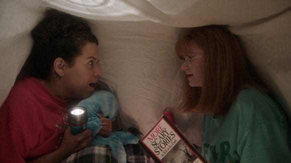 Two girls look at each other under a white blanket fort while holding a flashlight and a book.