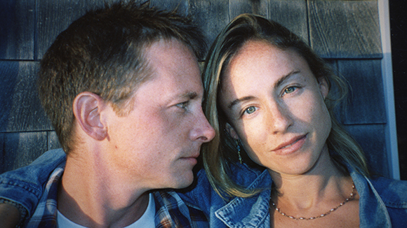 A man and a woman lean their heads together against a dark wooden shingle wall.