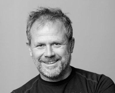 A smiling middle-aged man with short, messy hair and a beard looking toward the camera.