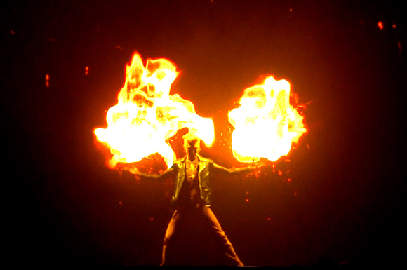 A person stands with arms outstretched between two large, bright bursts of orange and yellow flames.