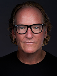 A man with wavy brown hair and black-rimmed glasses looking directly at the camera against a dark background.