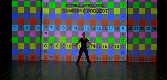 A person stands centered before a large, brightly lit grid of multicolored squares and numbers.
