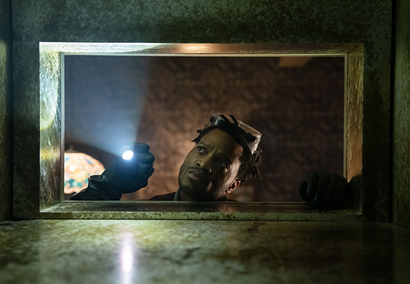 A man in dark clothing looks through a small window, holding a bright flashlight in a dark room.