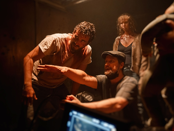 A man in a white shirt leans over a set while another man in a black shirt operates equipment.