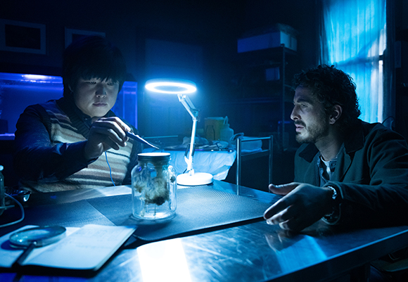 Two men lean over a table in a dark room illuminated by a blue ring light and a jar containing a specimen.