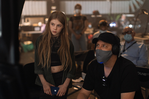 A young girl with long hair stands on a film set while a man in a mask looks on.