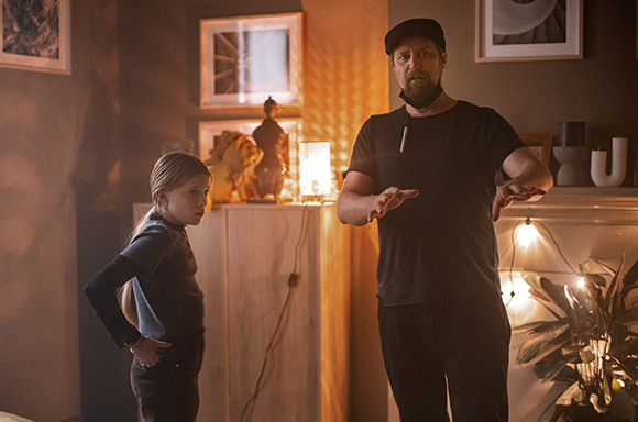 A man in a black cap and t-shirt gestures with his hands toward a young girl standing with hands on her hips.