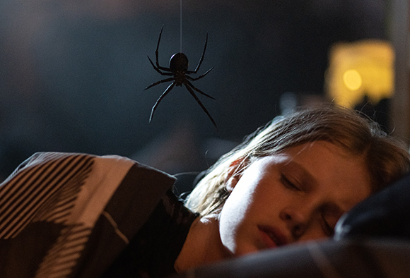 A black spider hangs from a thin thread directly above a young child sleeping in a dark room.