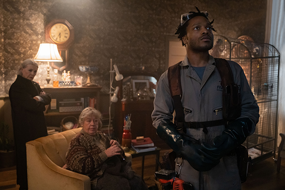 A man in a grey jumpsuit and heavy gloves looks upward in a dimly lit, cluttered living room.
