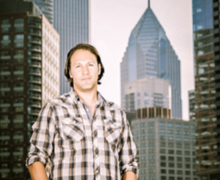 A man in a plaid shirt stands before a backdrop of tall city skyscrapers.