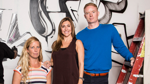A woman in a striped shirt, a woman in a dark tank top, and a man in a blue sweater stand before graffiti.