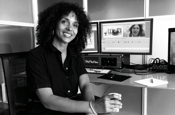A smiling woman with curly hair sits at a desk with multiple computer monitors in a black and white photo.