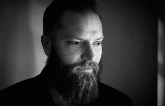 Black and white close-up portrait of a man with a thick beard looking slightly downward.