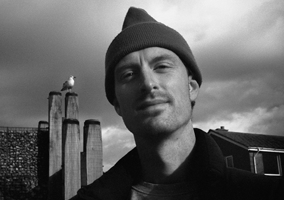 Black and white close-up of a man in a beanie with a seagull perched on a wooden post behind him.