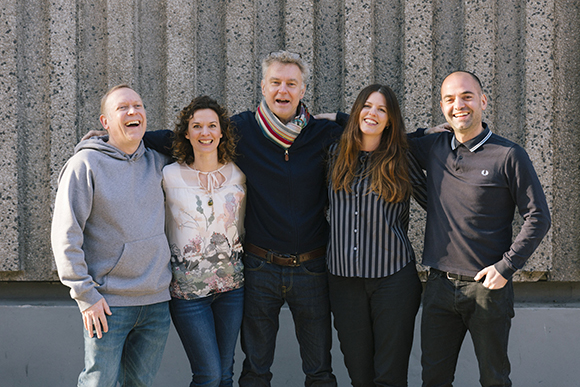 Five diverse adults smiling and posing together in front of a textured concrete wall.