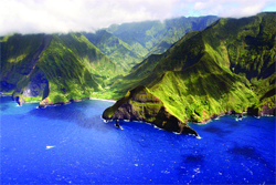 Aerial view of steep, green tropical mountains sloping into a bright blue ocean under a cloudy sky.