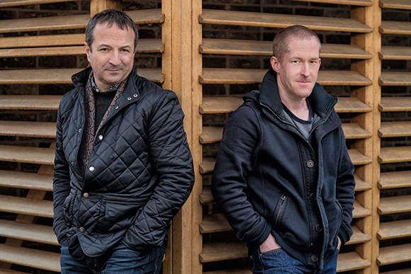 Two men in dark jackets stand back-to-back in front of a light brown wooden slatted wall.