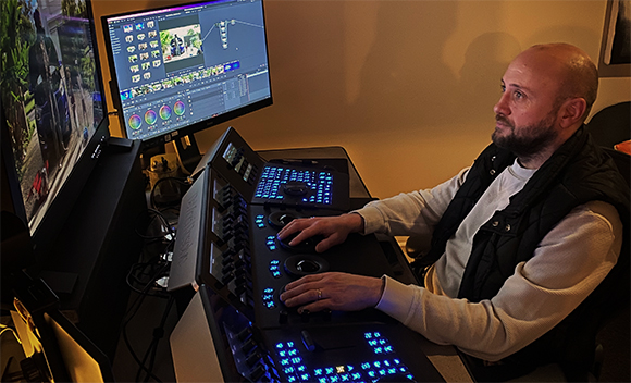 A man sits at a desk using a large control surface and dual monitors to edit video content.