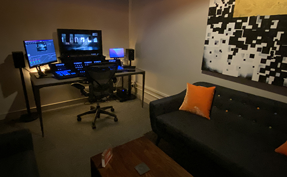 A production workstation with multiple monitors and a black desk next to a dark sofa with an orange pillow.