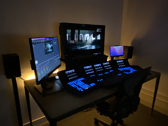 A dark workspace featuring a large monitor, a color grading panel, and a computer monitor on a desk.