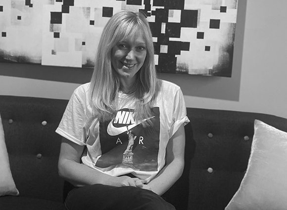 A smiling woman with long blonde hair wearing a Nike graphic t-shirt, sitting on a dark sofa.