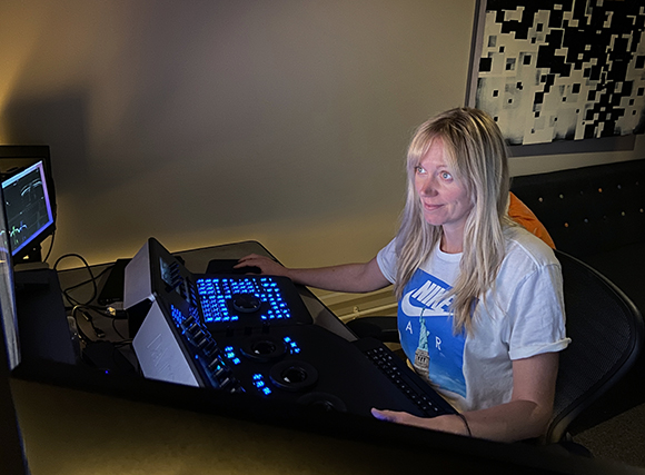 A blonde woman in a white Nike t-shirt operates a glowing blue control console in a dim studio.