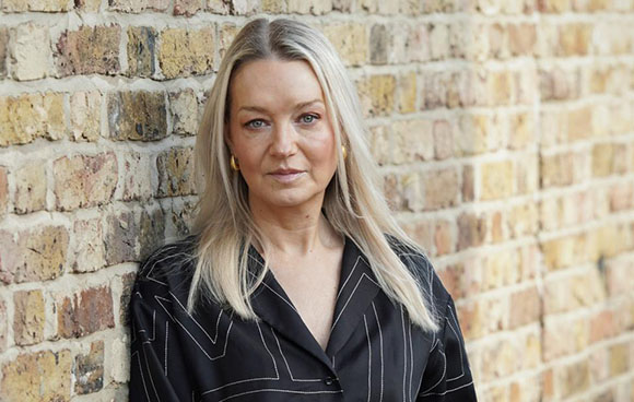 A woman with long blonde hair wearing a black patterned shirt leans against a brick wall.