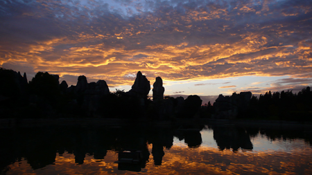 Silhouette of jagged rock formations and trees against a golden and purple sunset reflected in a calm lake.
