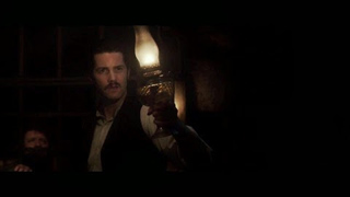 A man with a mustache holds up a glowing lantern in a dark, dimly lit room.