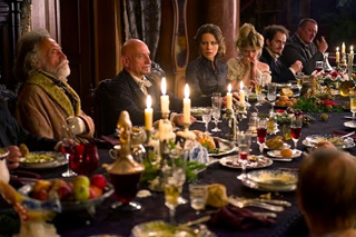 A group of people sit around a long dining table lit by many candles and filled with food and drink.