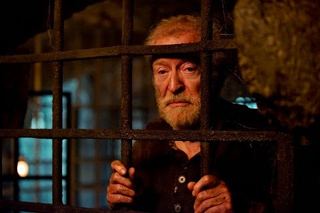 An elderly man with a beard looks out from behind dark, rusted metal prison bars.