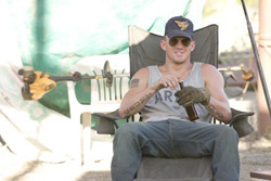 A man with sunglasses and a baseball cap sits in a folding chair holding a bottle.