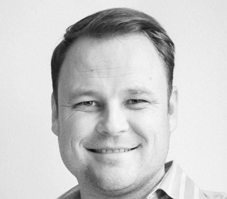 Black and white headshot of a smiling man wearing a striped shirt.