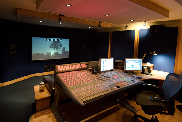 A large audio mixing console sits in a dark recording studio with monitors and acoustic panels.