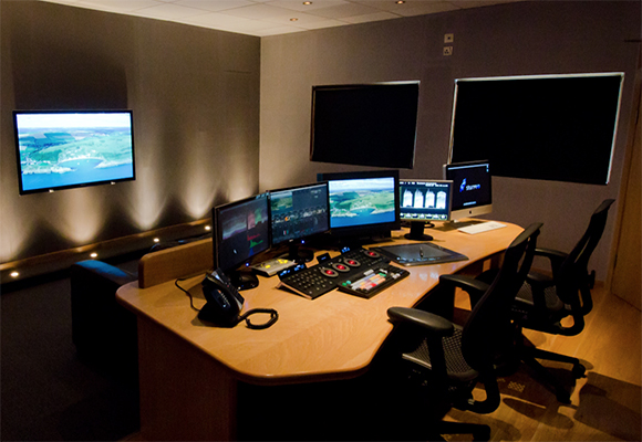 A control room featuring a large wooden desk with several computer monitors, keyboards, and black office chairs.