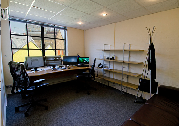 A small office with a long wooden desk, two computer monitors, black office chairs, and wall-mounted shelving.