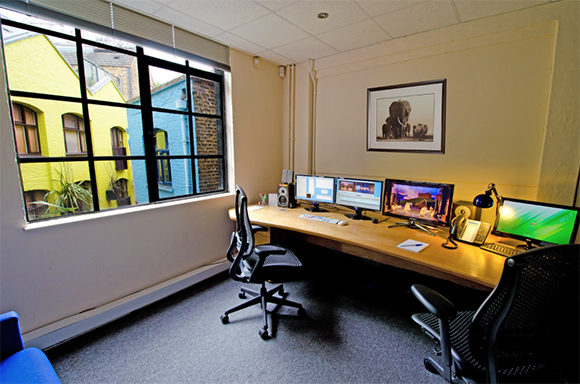 Office workspace with a long wooden desk, four computer monitors, black ergonomic chairs, and a window view.